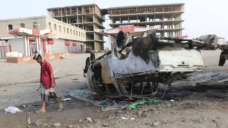 An armed man walks next to a destroyed vehicle at the site of a deadly car bomb attack in the port city of Aden, Yemen. © AP / Wail al-Qubaty