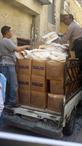 A small UNRWA van delivers boxes of staple foods to Yarmouk camp residents who wait at a pick-up point. Bread donated by the Syrian government lies atop the boxes.