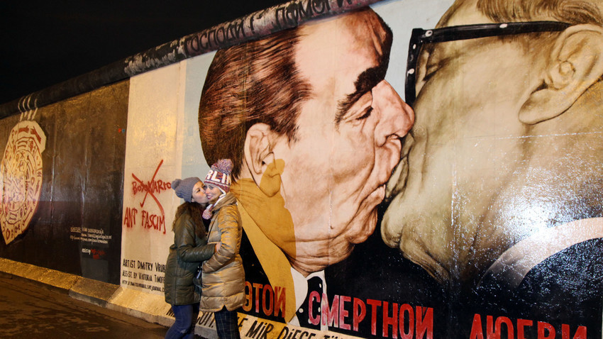 Dua orang wanita berpose di depan lukisan grafiti karya Dmitri Vrubel yang menggambarkan ciuman persaudaraan antara Pemimpin Soviet Leonid Brezhnev (kiri) dan Pemimpin Jerman Timur Erich Honecker di East Side Gallery, Berlin Wall art, Jerman.