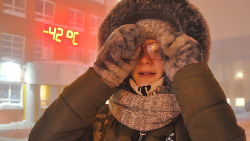 Seorang perempuan berdiri berlatarkan papan informasi suhu yang menunjukan -42 °C di Norilsk. Denis Kozhevnikov