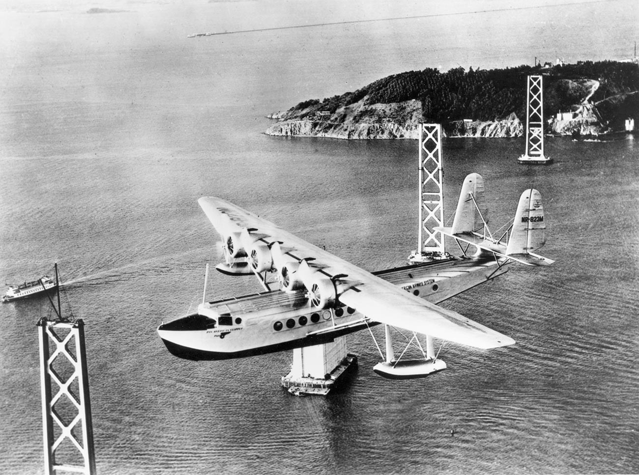 Sikorsky S-42 „Pan American Clipper“ прелеће залив Сан Франциска на путу за Хаваје. Види се конструкција моста Сан Франциско – Окланд у изградњи. Око 1934.
