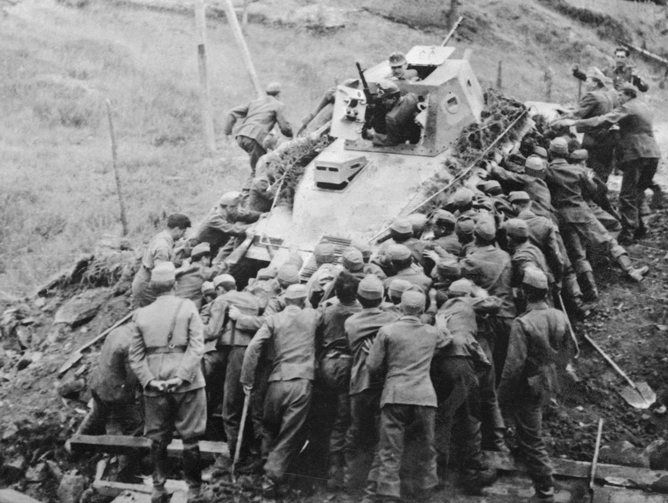 Over 50 Hungarian soldiers are putting their shoulders to the tractor wheels in an effort to recover this disabled Soviet tank.