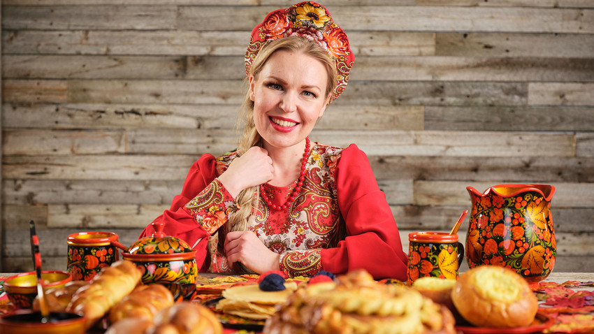 Жена у традиционалној руској ношњи са кокошником на глави за трпезом на којој се налази празнични хлеб.
