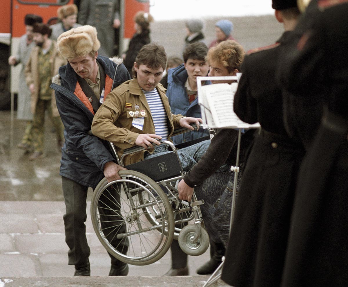 Several people helping a disabled vet of the war in Afghanistan to get up the stairs in his wheelchair, 1990.