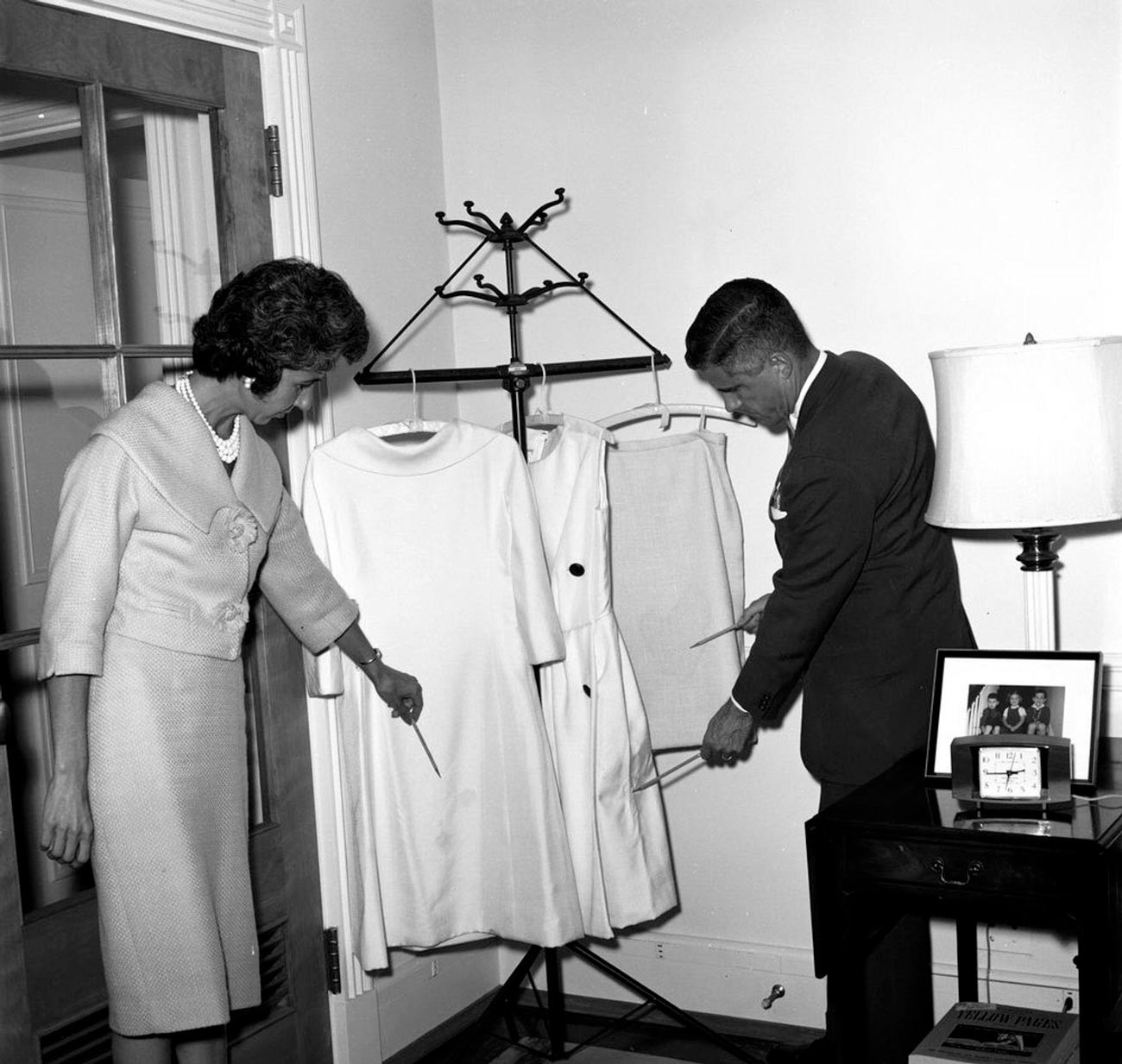 Robe pour Jacqueline Kennedy. La secrétaire de la première dame, Mary Gallagher, et un homme non identifié examinent deux robes et une jupe provenant d'un carton d'expédition endommagé envoyé par la société de création de mode d'Oleg Cassini à la Maison Blanche.