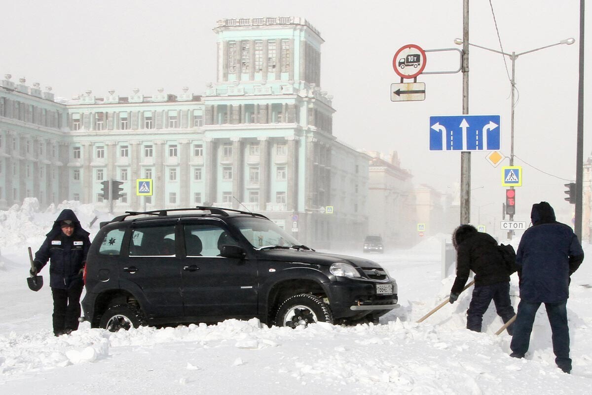 Domačini v Norilsku odkopavajo avto iz snega, medtem ko piha močan veter.