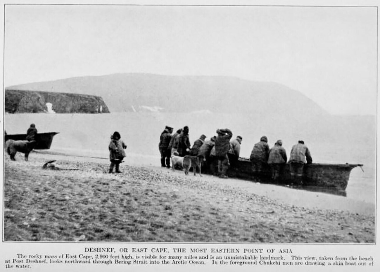 Orang-orang Chukchi di Port Dezhnev membawa umiak ke pantai; Tanjung Dezhnev di latar belakang.