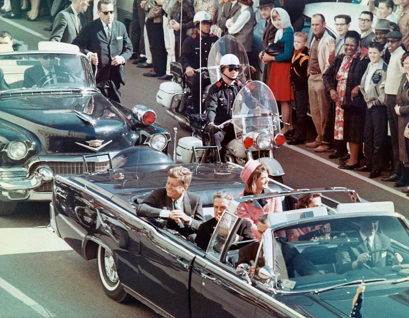 Presiden John F. Kennedy dan istrinya tersenyum pada kerumunan yang berbaris di rute iring-iringan mobil mereka di Dallas, Texas, pada 22 November 1963. Beberapa menit kemudian, Presiden dibunuh saat mobilnya melewati Dealey Plaza.