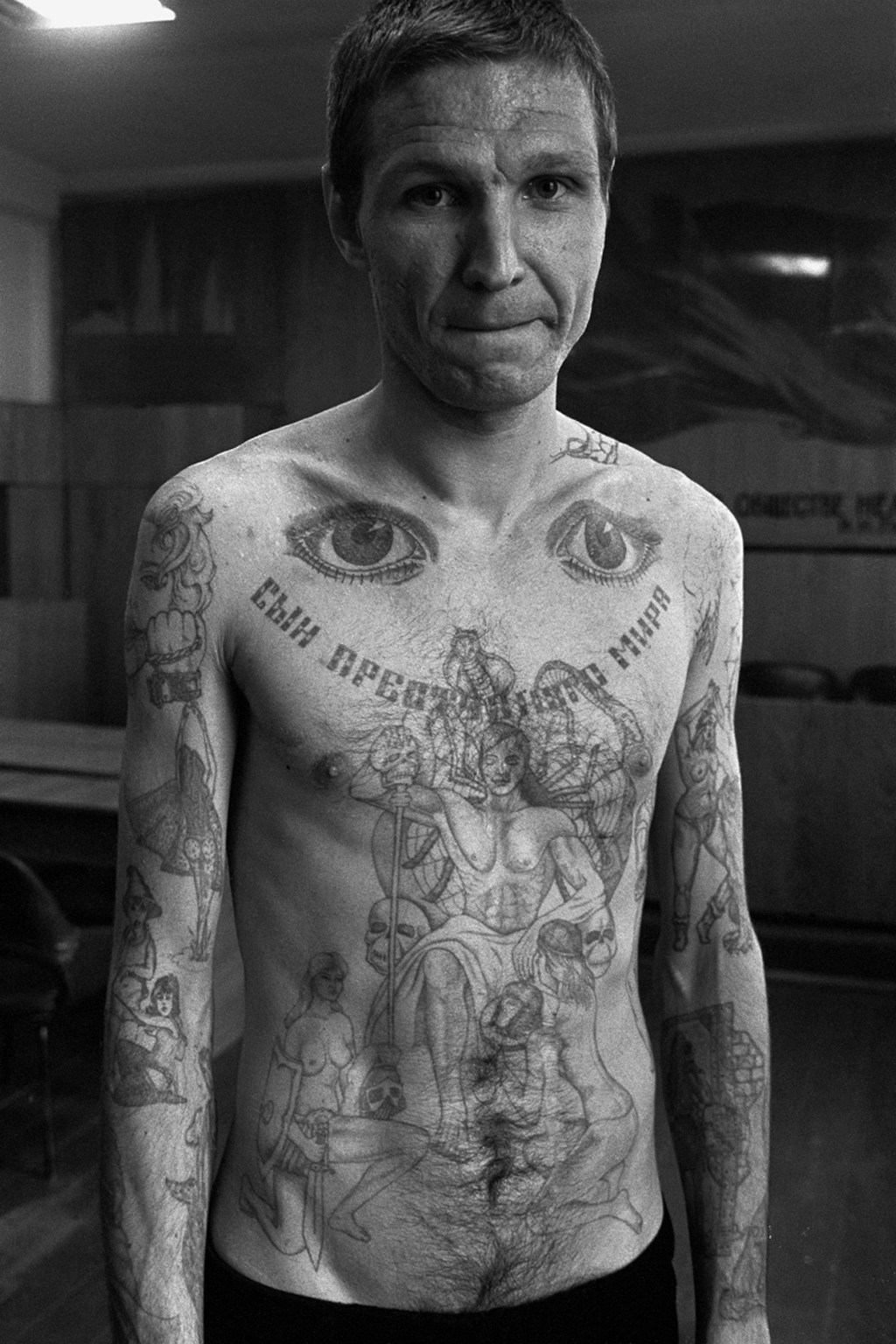 The eyes on the top of the chest signify 'I can see everything' and 'I am watching'. Text across the chest reads 'Son of the criminal world'. This photograph shows tattoos in a combination of old and new styles. In the ‘new’ style a large number of almost random images on the convict’s body. In the ‘traditional’ style there is one large central tattoo on the chest, filling as much space as possible.