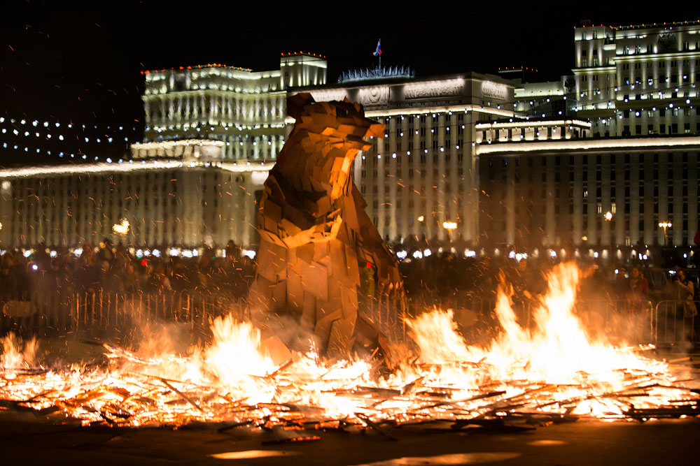 People watch the burning symbol of spring, which looks like a brown bear getting out of his den, created by Hungarian artist Gabor Miklós Szoke for the festival in Gorky Park during a celebration of Maslenitsa, or Shrovetide in Moscow, Russia, Sunday, March 13, 2016, with the building of the National Defense Center of Russian Defense Ministry in the background. Maslenitsa is a traditional Russian holiday marking the end of winter that dates back to pagan times.