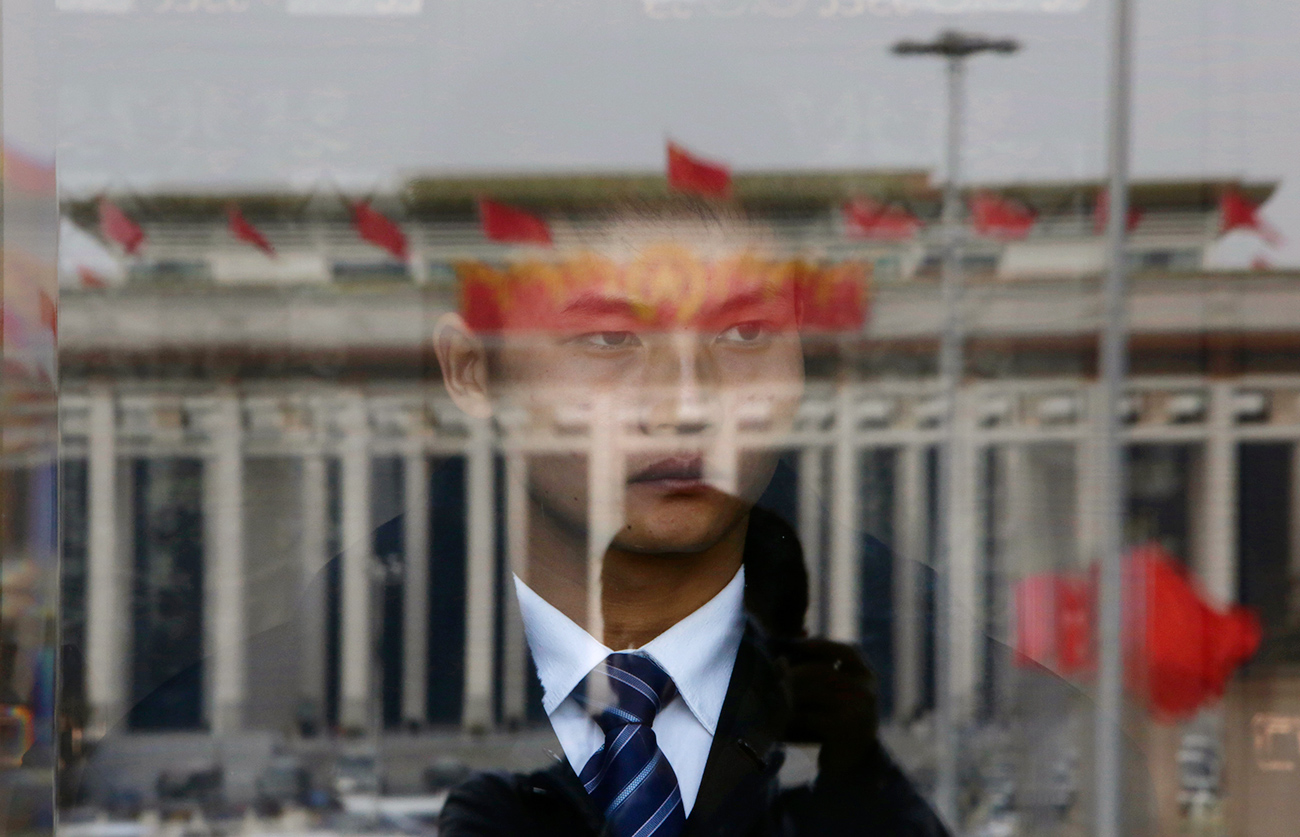 A soldier in plain clothes from the Chinese People's Liberation Army (PLA) stands guard behind an entrance of the Great Hall of the People, the venue of the annual session of China's parliament, as red flags are reflected on a glass door in Beijing