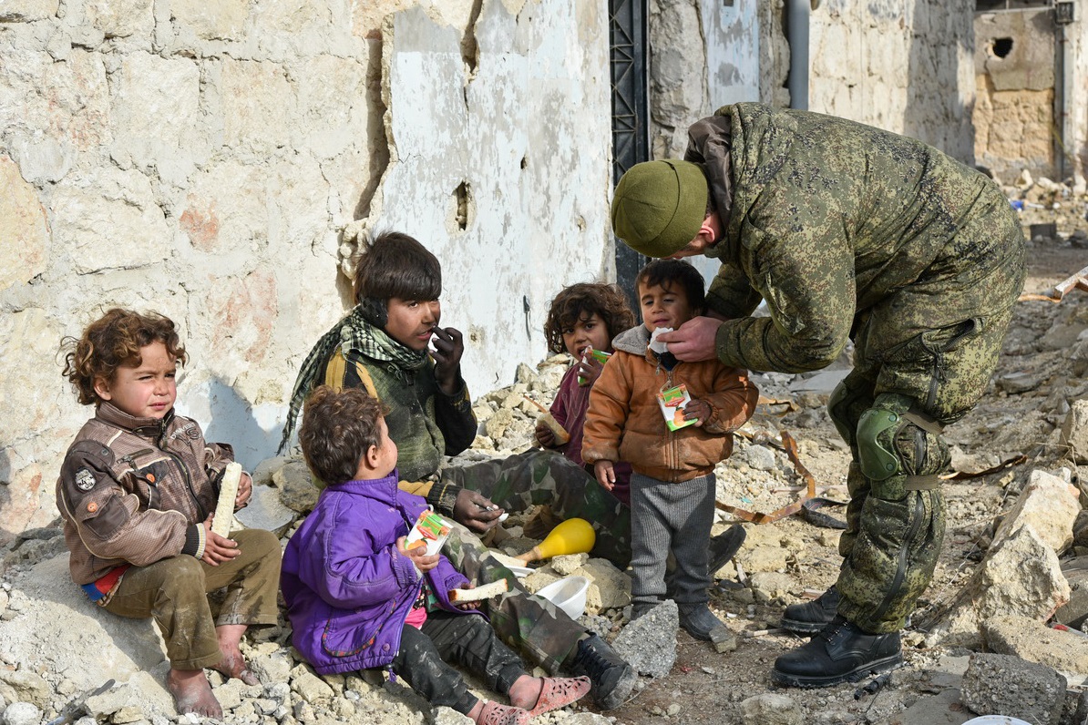 Seorang tentara Rusia tengah membersihkan mulut seorang anak Suriah di antara teman-temannya yang duduk di sekitar puing-puing bangunan.